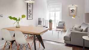 Un salon moderne avec un coin repas, comprenant une table en bois, des chaises blanches, un canapé gris et une grande fenêtre.