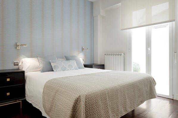 A bedroom with a large bed, a nightstand, a wall-mounted light, and a window with a view.