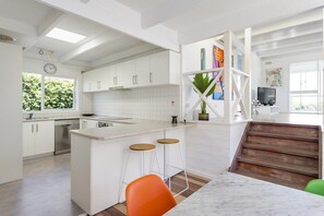 A modern kitchen with white cabinetry, a central island with stools, a dining area with a table, and a staircase leading to an upper level.