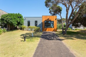 A modern house with a paved driveway leading to a wooden door, surrounded by a well-maintained lawn and trees. - At Agnes Ave Blairgowrie (Blairgowrie)