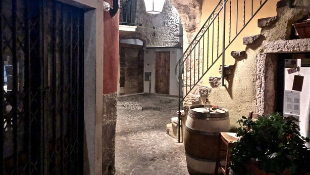 A narrow cobblestone alleyway with a rustic stone wall, a wooden barrel, and a metal railing. - Apartment del Pescatore In Downtown (Torri del Benaco)