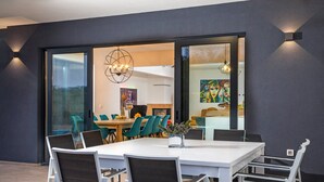 Een moderne eetruimte met een witte tafel en stoelen, een kroonluchter en uitzicht door glazen deuren op een keuken en eetgedeelte.