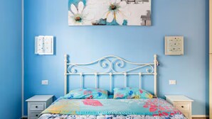 A bedroom with a blue wall, a white metal bed frame, a floral bedspread, and a painting of flowers on the wall.