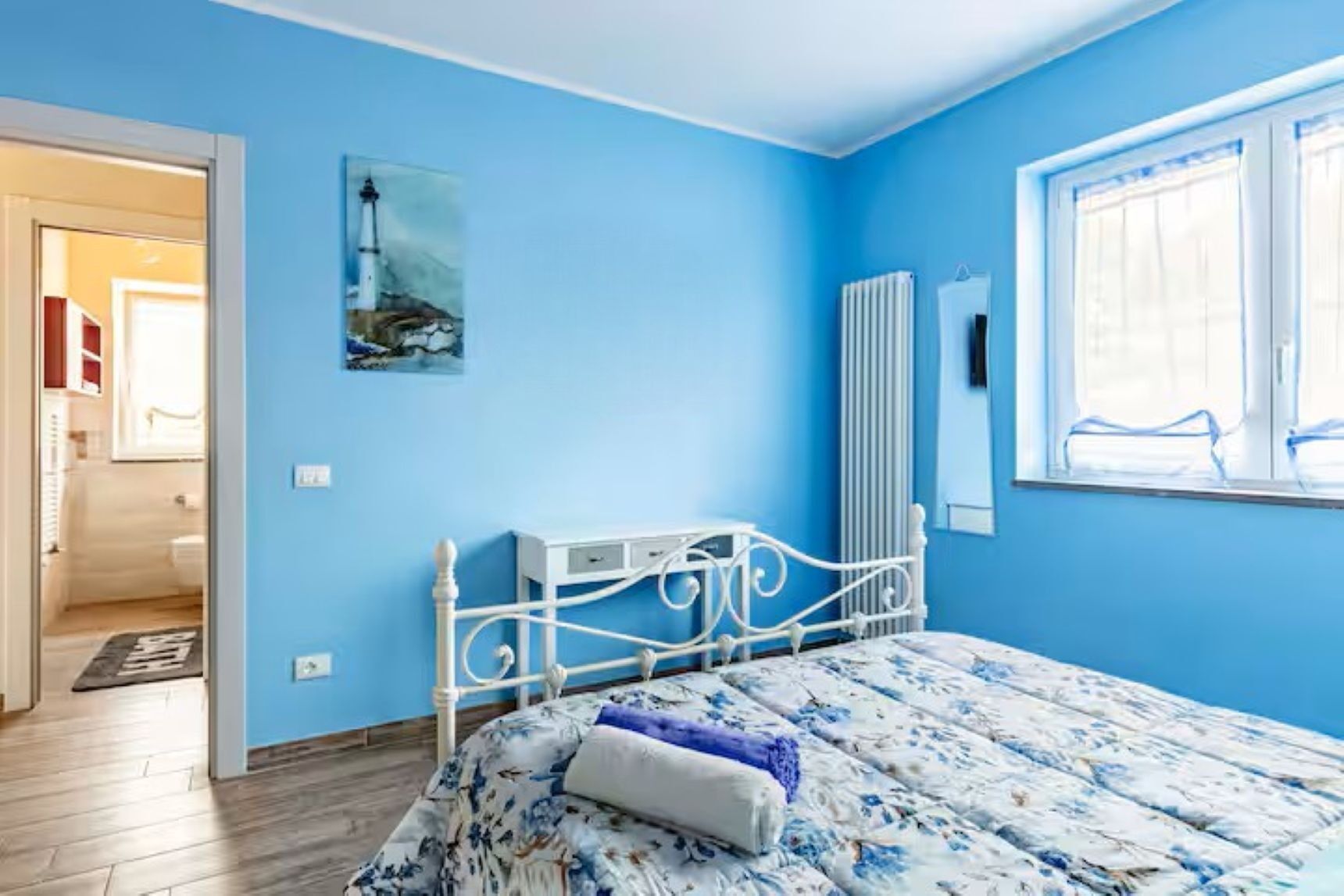 A bedroom with a bed, a radiator, a window, and a door leading to another room.