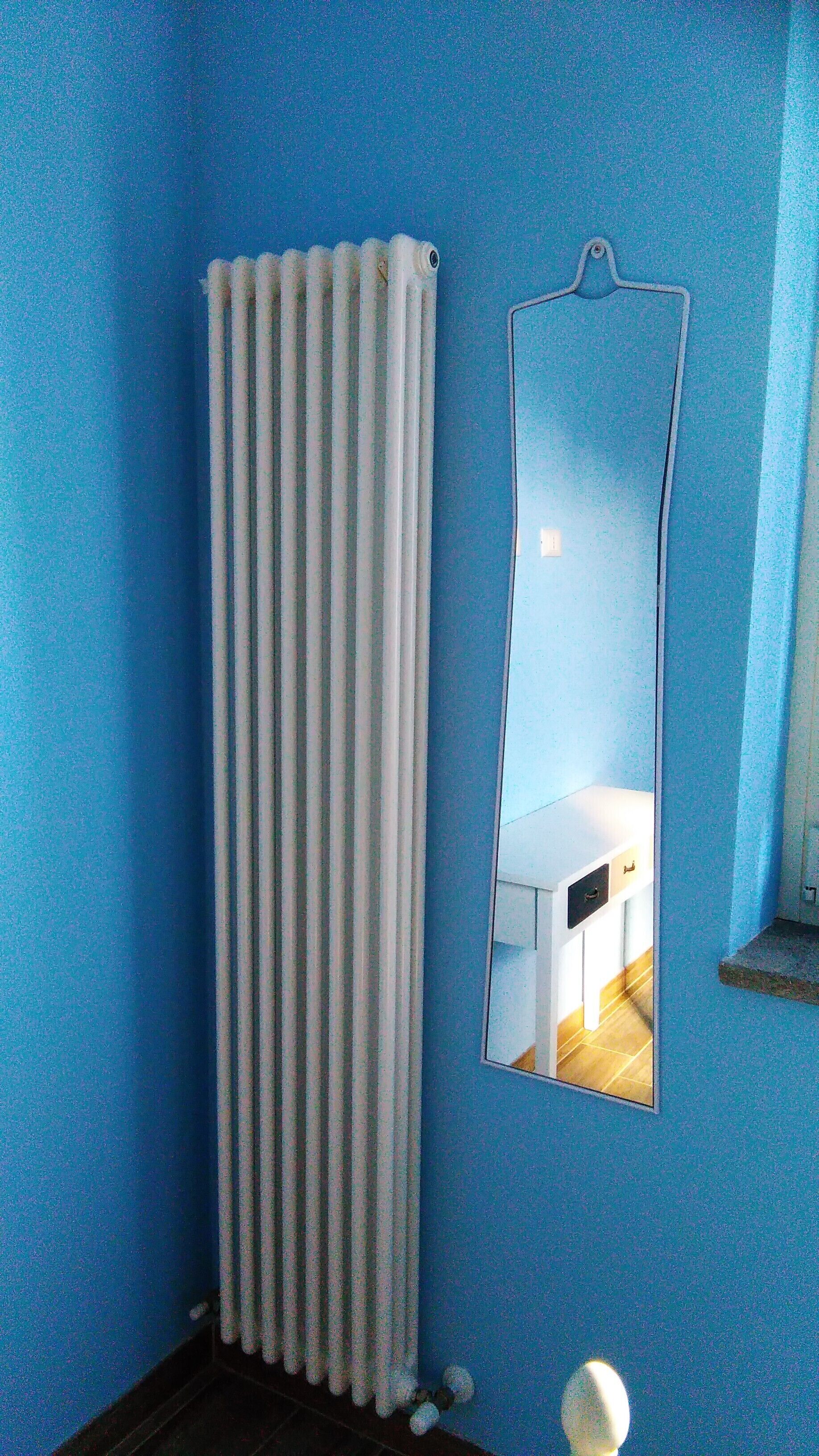 Un bagno con una parete blu, un termosifone bianco e uno specchio che riflette una stanza con un letto e una finestra.