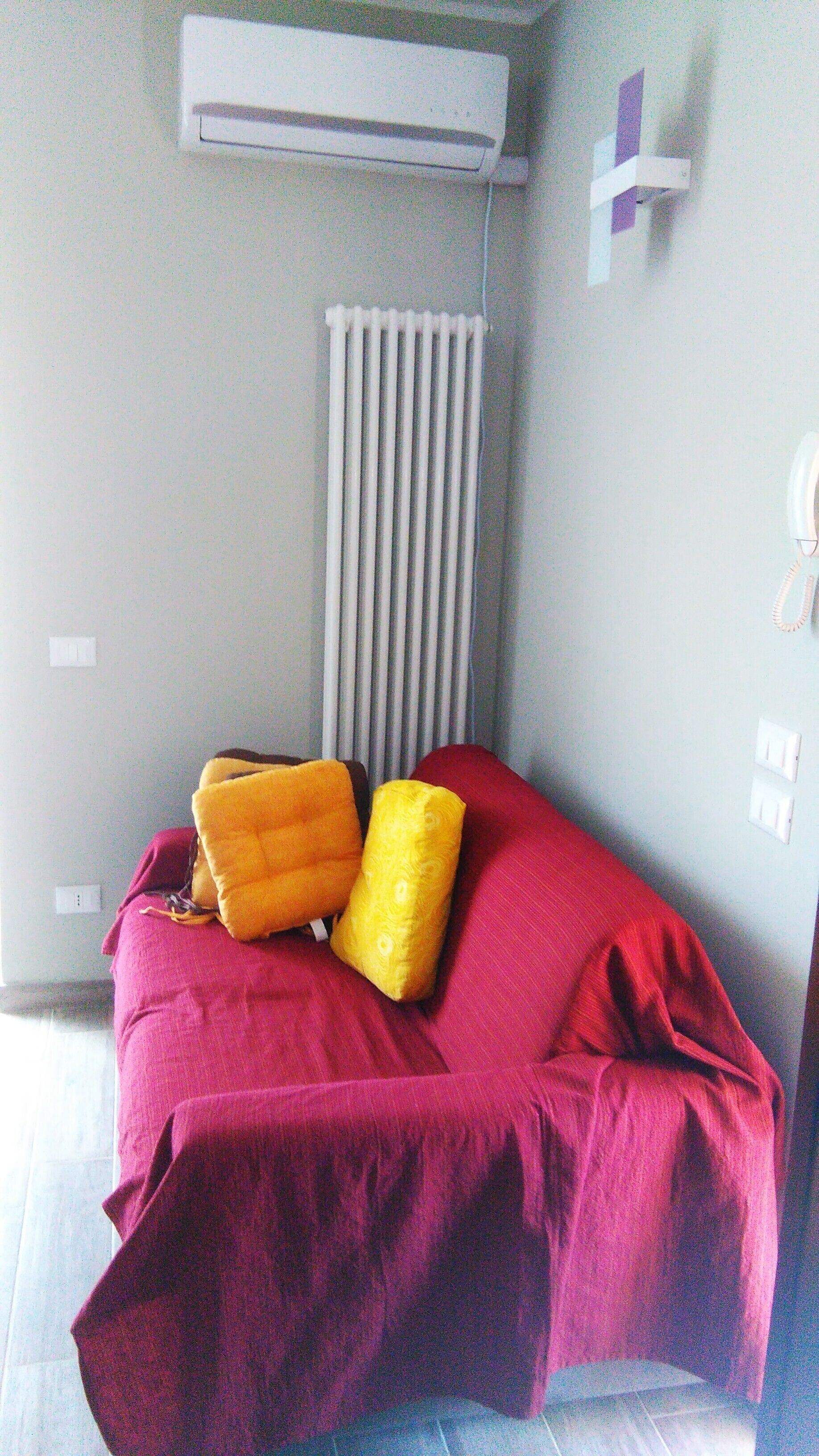 Une chambre avec un lit recouvert d’une couverture rouge, deux oreillers, un appareil de climatisation blanc placé au-dessus du lit, un radiateur et un téléphone fixé au mur.