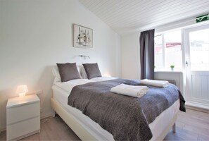 A neatly made bed with a grey blanket and pillows, a bedside lamp, and a small table with a lamp.