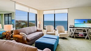 A modern hotel room with a large window offering a view of the ocean, a flat-screen TV, a sofa, armchairs, and a desk with a lamp. - SS 81122 Expansive Ocean & Intracoastal Waterway View Condo - Vacation Rental in Ocean Village (Fort Pierce)