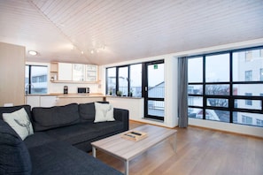 A modern living room with a grey sectional sofa, a wooden coffee table, and a kitchen area with cabinets and a microwave. - Penthouse Apartment - Skolavordustigur (Reykjavík)
