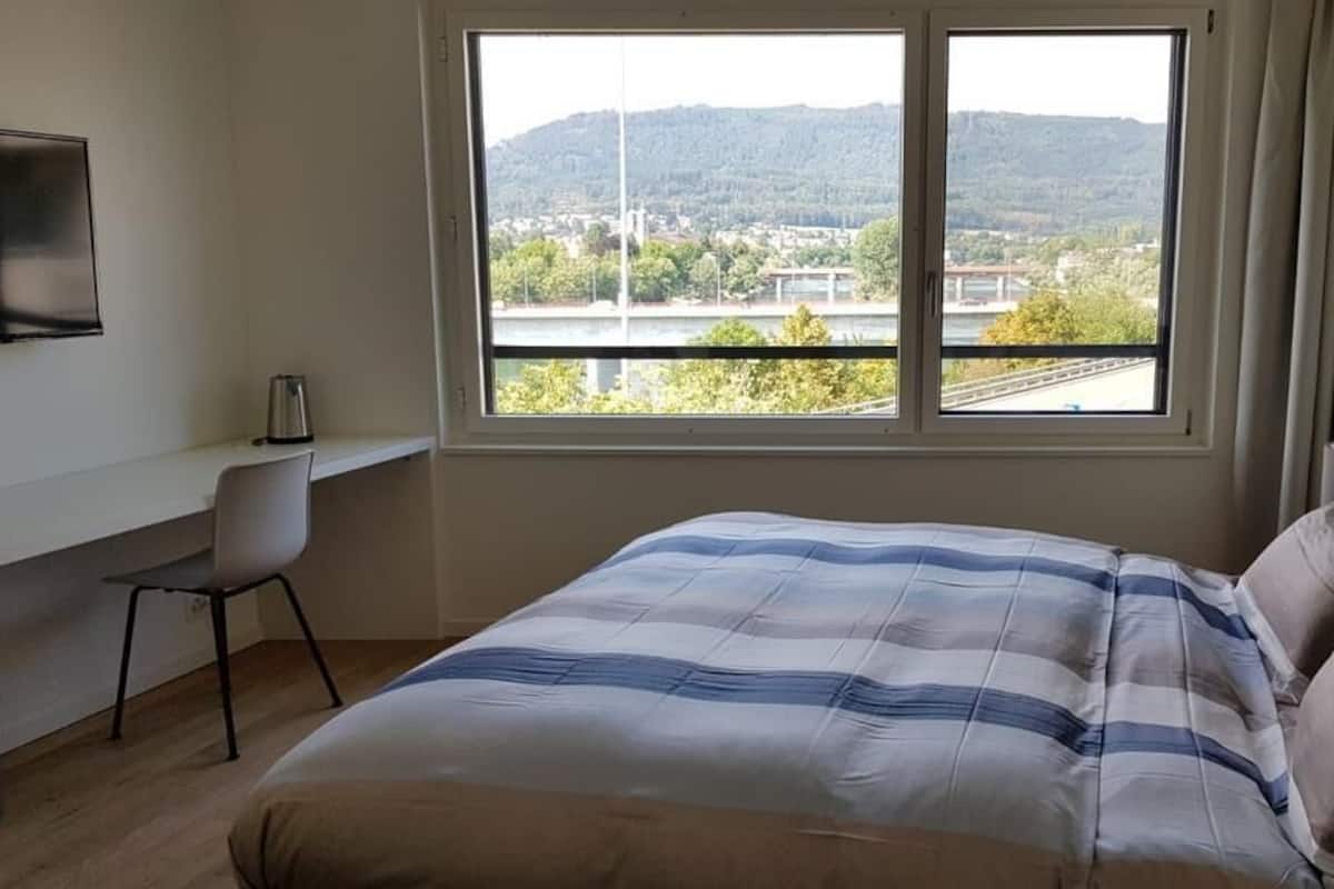 Ein Hotelzimmer mit Bett, Schreibtisch, Stuhl und großem Fenster mit Blick auf eine Brücke und Hügel.