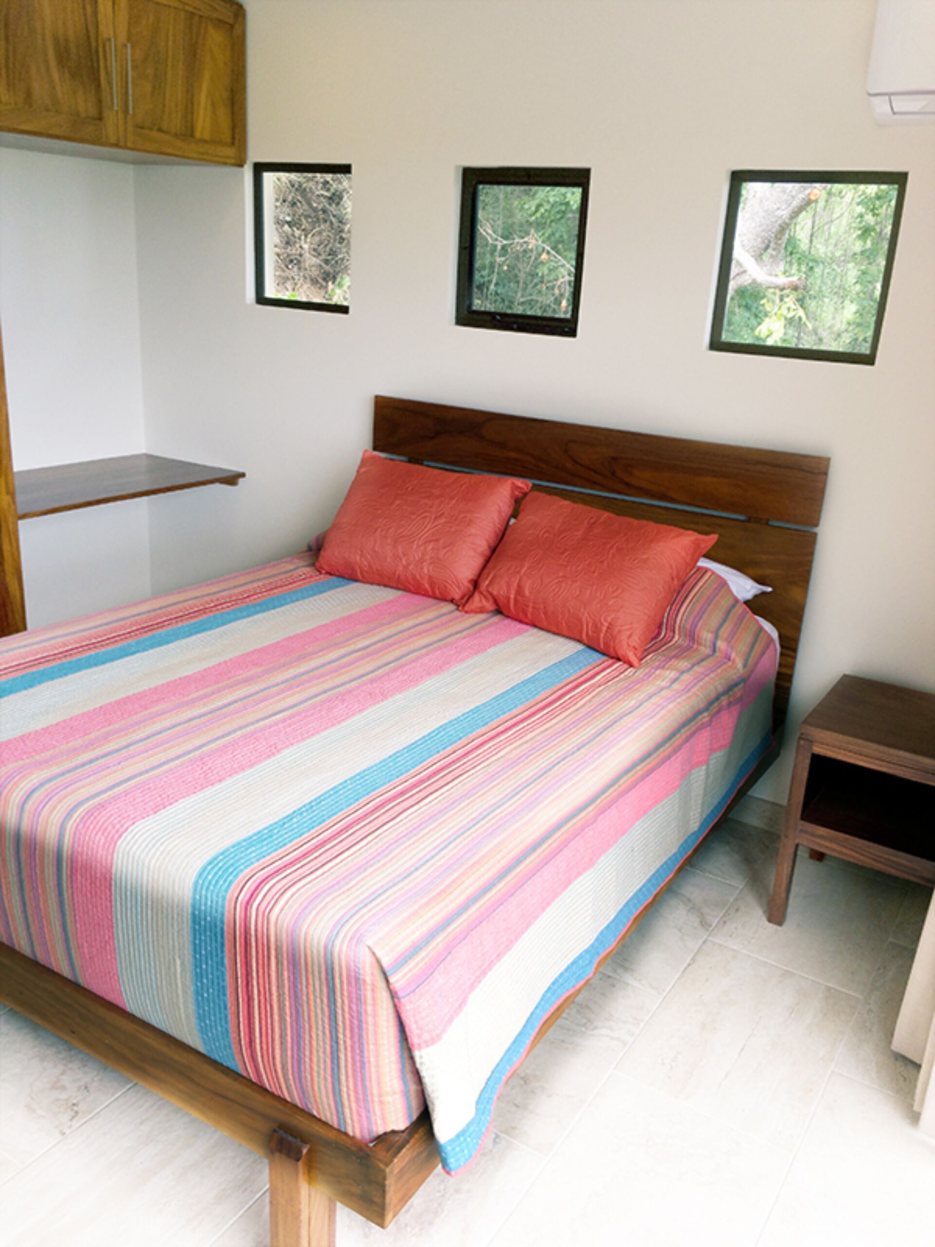 A bed with a colorful striped bedspread, wooden headboard, and two pillows. There are framed pictures on the wall and a small wooden nightstand beside the bed.
