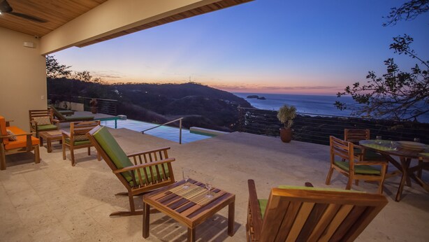 A wooden deck with outdoor furniture, a pool, and a view of the ocean at dusk. - Casa de la Paz (Sardinal)
