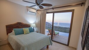 A bedroom with a bed, a ceiling fan, a view of the ocean through a sliding door, and a balcony with a chair. - Casa de la Paz (Sardinal)
