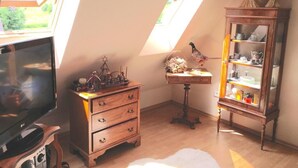 Ein Zimmer mit schrägem Dach, einem Fernseher, einer Holzkommode, einem kleinen Tisch mit Lampe und einer Vitrine.