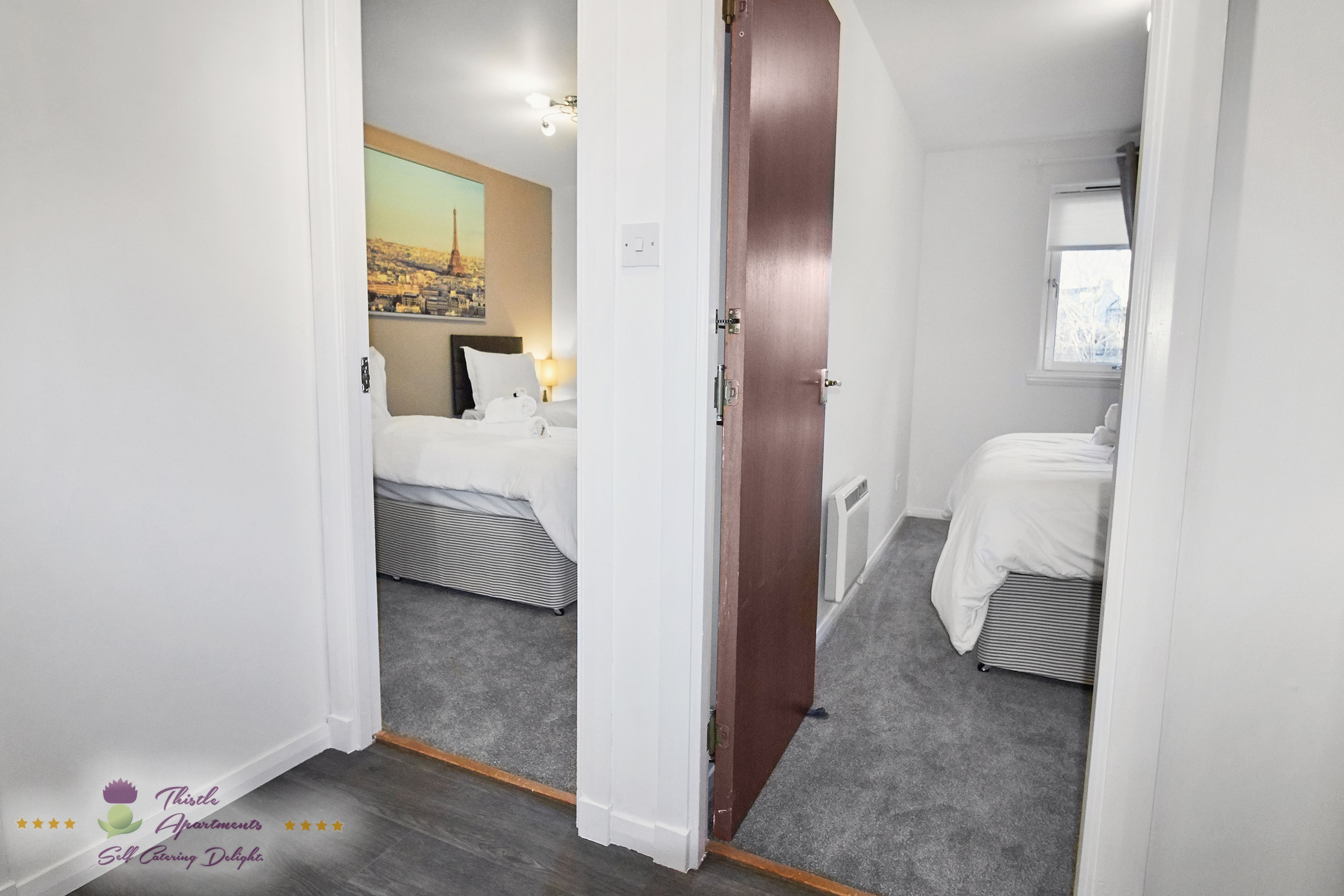 A narrow hallway leading to two bedrooms with separate beds, a window, and a cityscape painting on the wall.