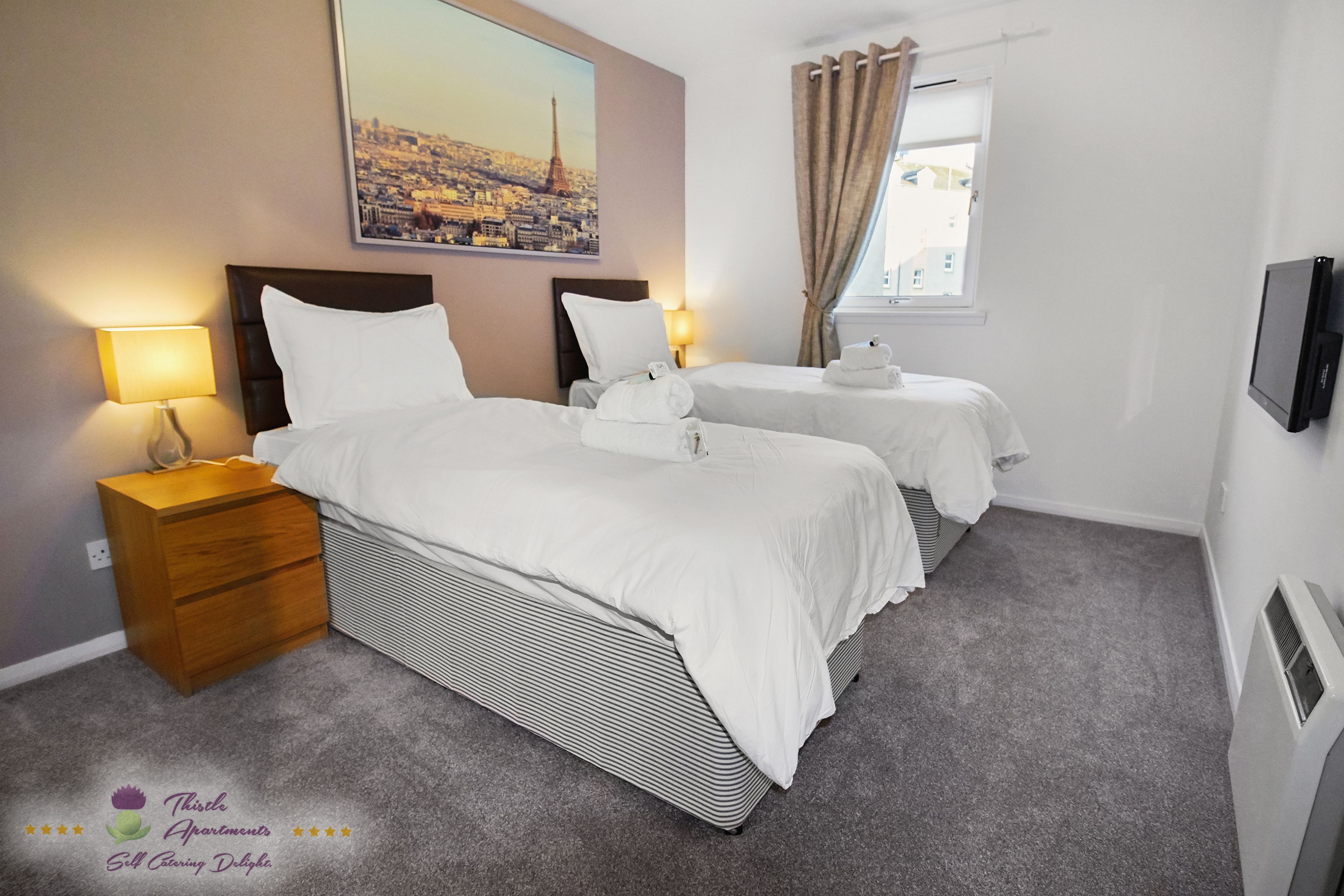 A hotel room with two beds, a nightstand, a lamp, a TV, and a window with curtains.