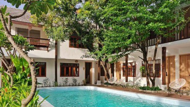 Un área de piscina con una piscina azul cristalina, rodeada de exuberante vegetación y un edificio de dos pisos con balcones.
