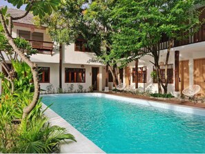 Un área de piscina con una piscina azul cristalina, rodeada de exuberante vegetación y un edificio de dos pisos con balcones.