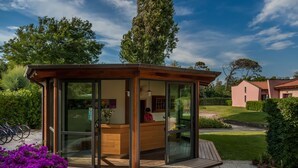 A modern, glass-enclosed reception area with a wooden deck, surrounded by lush greenery and a clear sky. - Miramare Resort (Pisa)