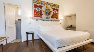 A hotel room with a bed, a small table, a chair, a wall-mounted lamp, and a decorative tapestry.