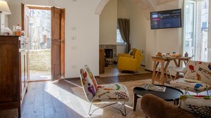 A cozy living room with a wooden cabinet, a yellow armchair, a patterned ottoman, and a television mounted on the wall. - L' Artiere Dimore nei Sassi (Matera)
