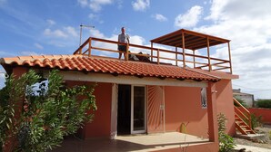 A two-story building with a red-tiled roof, a balcony on the upper floor, and a door leading to an enclosed patio area. - Full equipped bungalows with swimming pool, wifi, garden for 2 to 5 people (Morro)