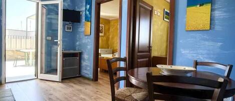 Uma sala de jantar com mesa redonda e cadeiras, porta de vidro deslizante que dá para uma varanda, e área de cozinha visível através da porta.