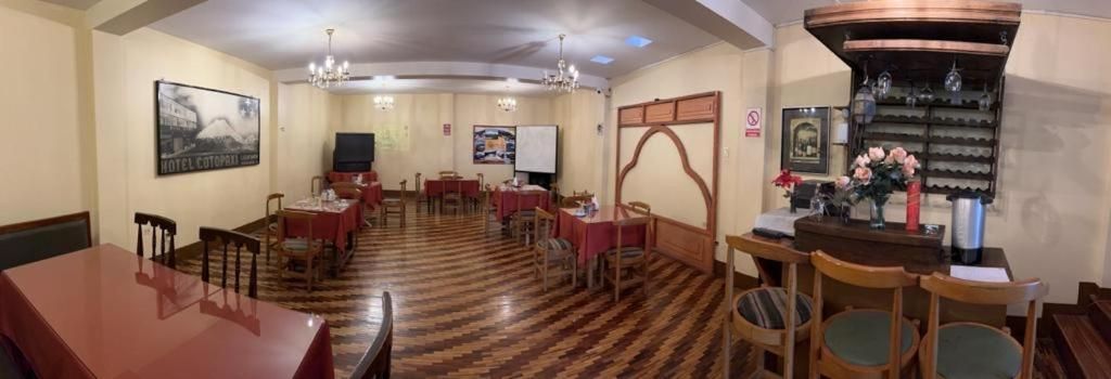 Un comedor de hotel con varias mesas y sillas, una zona de bar con mostrador y estanterías, y una pantalla grande en la pared.