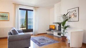 Una sala de estar moderna con un sofá gris, un sillón blanco, un ventanal con vistas a montañas, una planta en maceta y una chimenea.