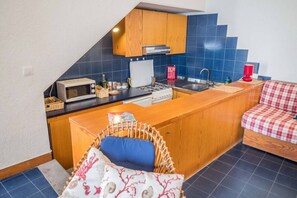 A compact kitchen with a microwave, stove, and sink. There's a wicker chair with a cushion and a checkered sofa.