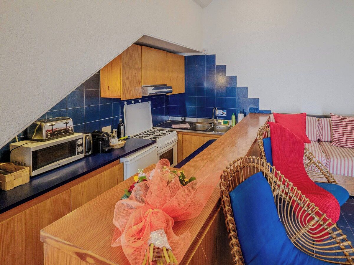 Une cuisine compacte équipée d’un four à micro-ondes, d’un grille-pain et d’une cuisinière, jouxtant un coin repas avec une table en bois et des coussins bleus.