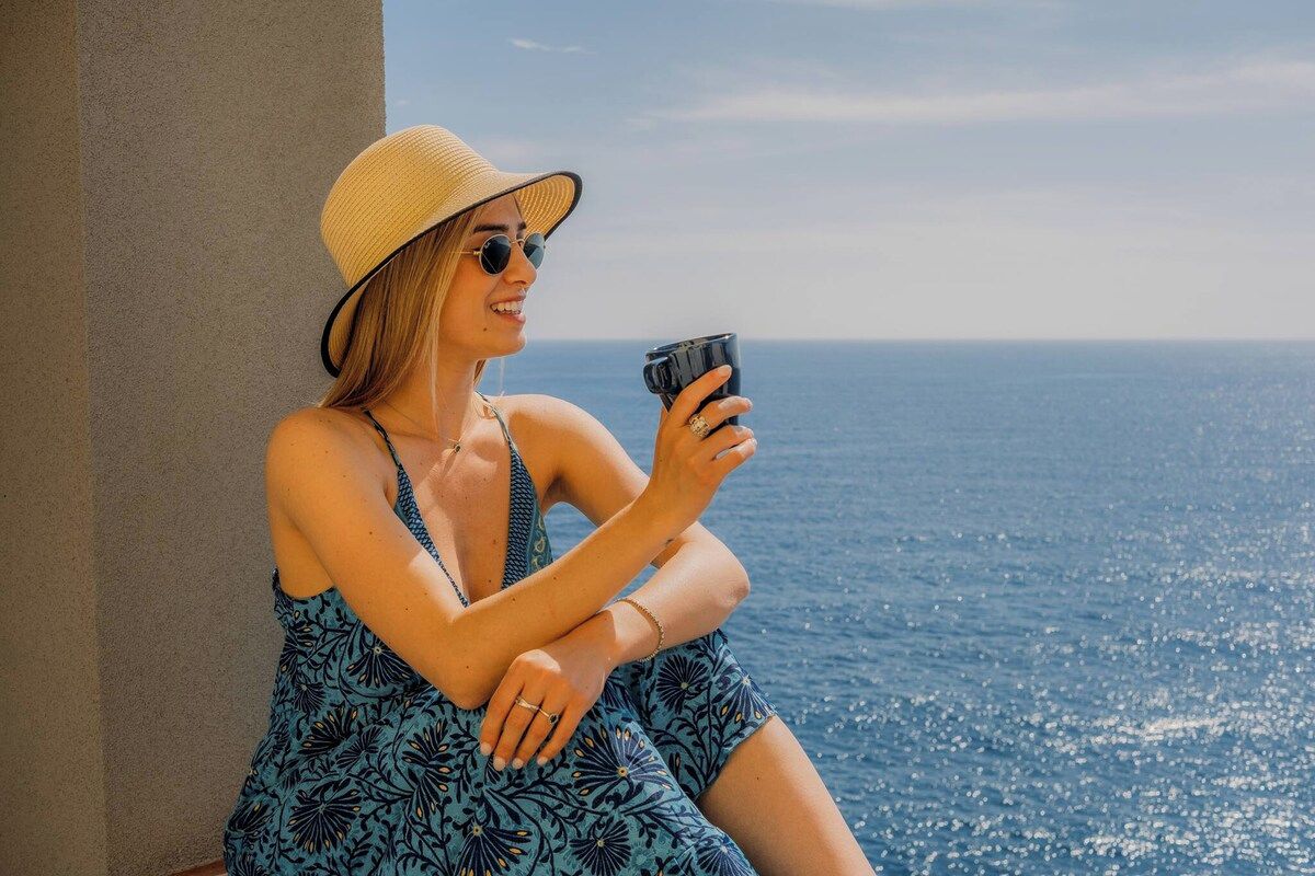 A person seated by a window overlooking a body of water, holding a mug.