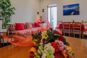 Sala de estar aconchegante com sofá, mesa de jantar e cadeiras. Uma grande janela oferece vista para o mar.