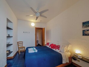 Ein Schlafzimmer mit einem Bett, einem Sessel, einem Deckenventilator, einem Nachttisch mit Lampe und einer Tür zu einem anderen Raum.
