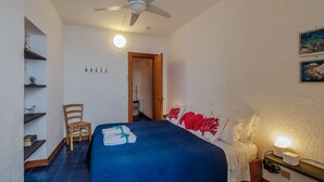 Ein Schlafzimmer mit Bett, Stuhl, Deckenventilator, Nachttisch mit Lampe und einer Bücherablage.