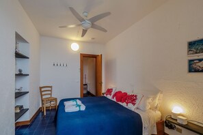Een slaapkamer met een bed, een stoel, een plafondventilator, een nachtkastje met een lamp en een plank met boeken.