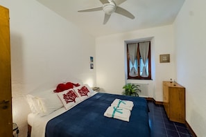 Quarto de hotel com cama, ventoinha no teto, janela com cortinas, radiador e um armário de madeira.