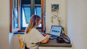 Eine Person arbeitet an einem Schreibtisch neben einem Fenster mit Jalousien, auf dem Tisch stehen ein Laptop und eine Kamera.