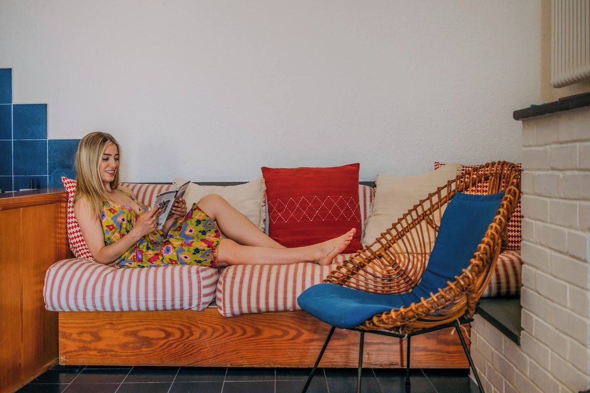 A person sitting on a sofa with striped cushions, a red pillow, and a blue chair with a woven backrest.