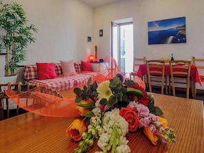Una habitación con una mesa de comedor dispuesta para dos personas, un sofá con funda a cuadros, una silla con cojín rojo y un cuadro colgado en la pared.