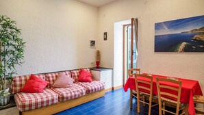 Ein gemütliches Zimmer mit einer karierten Couch, einem Esstisch mit rotem Tischläufer und einem großen Fenster mit Blick auf die Küste.