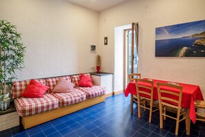 Une pièce chaleureuse avec un canapé à carreaux, une table à manger recouverte d’une nappe rouge et une grande fenêtre donnant sur la côte.