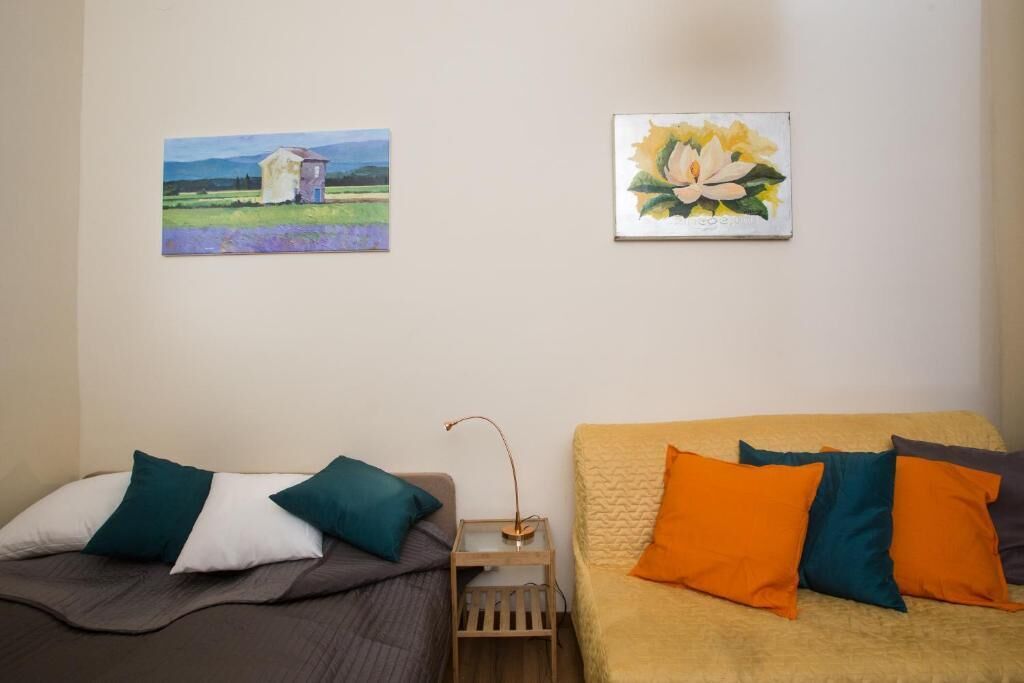 A bedroom with two beds, one with teal and white pillows and the other with orange and teal pillows. There are two paintings on the wall, one of a house and the other of a flower.