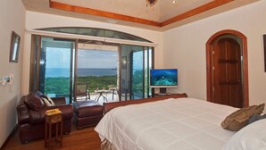 A bedroom with a large bed, a TV, a ceiling fan, and a view of the ocean. - Casa de los Suenos (Sardinal)