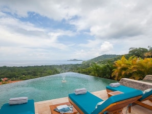 An infinity pool with a view of the sea and hills, surrounded by lounge chairs and a stone wall.