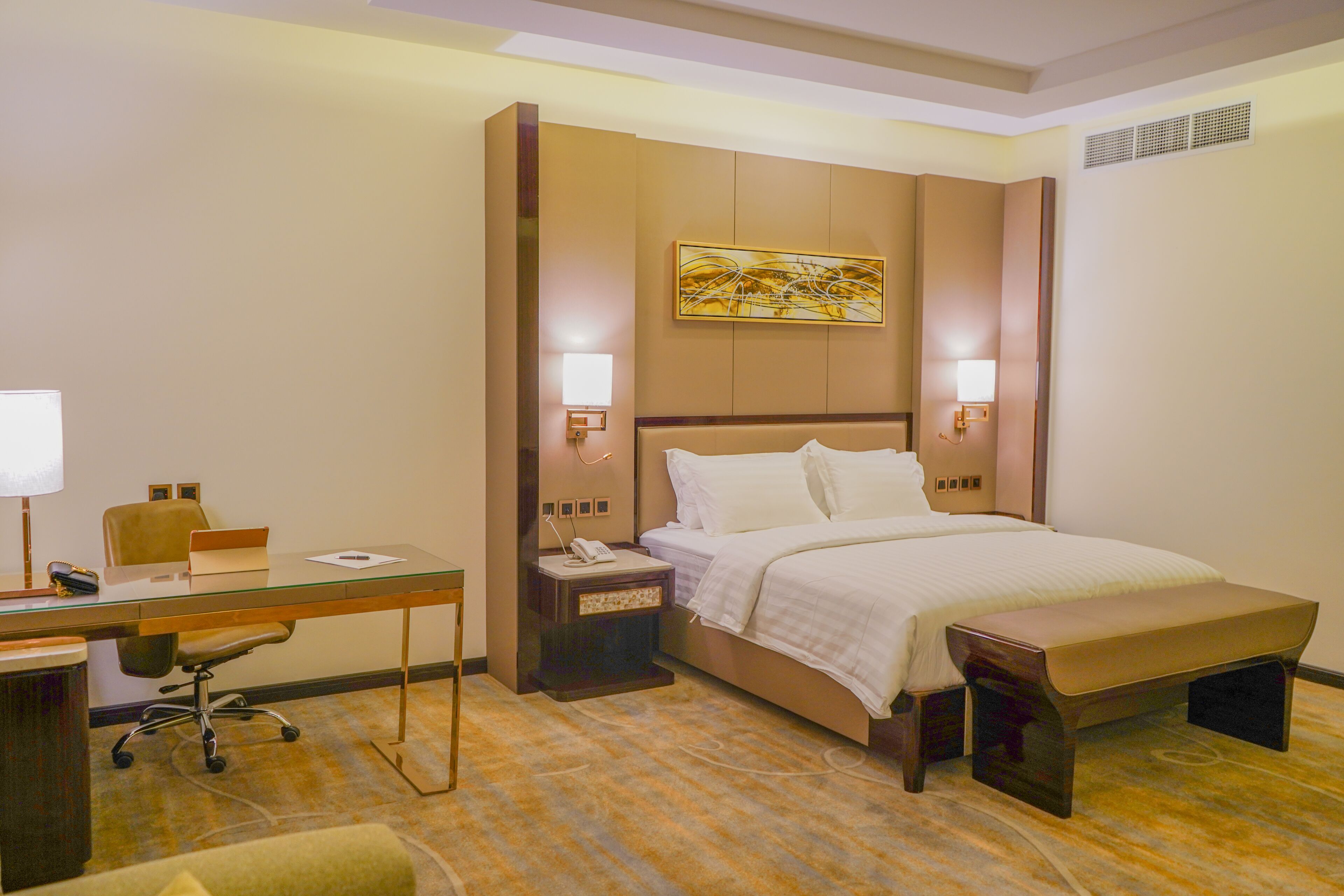 A hotel room with a bed, desk, chair, bedside tables, and artwork on the wall.