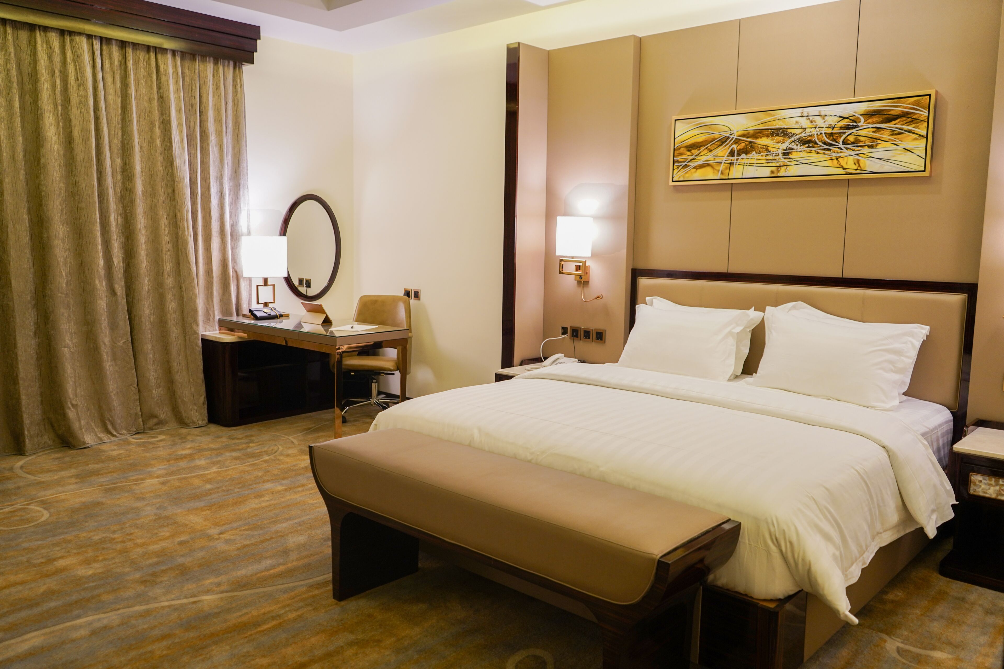 Habitación de hotel con una cama grande, un escritorio con silla, un banco, una mesita de noche, una lámpara, un espejo y un cuadro en la pared.
