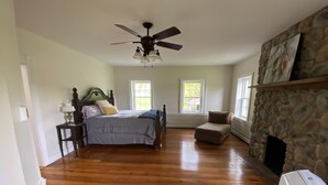 Ein geräumiges Schlafzimmer mit einem großen Bett, einem Deckenventilator, einem Kamin aus Stein und zwei Fenstern für natürliches Licht.