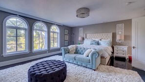 A bedroom with a large bed, a tufted sofa, and a round ottoman. There are three large windows and a chandelier.
