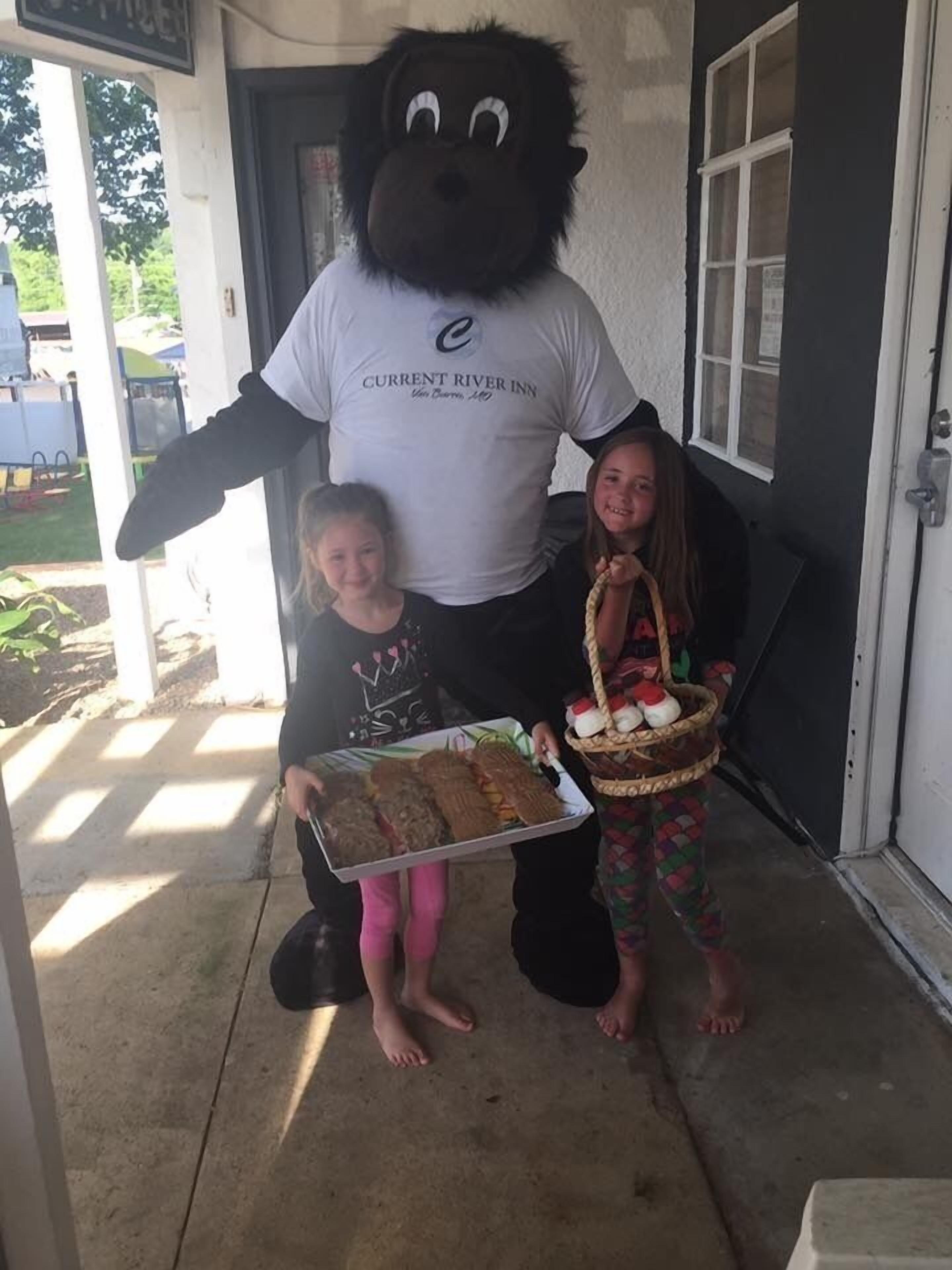A mascot holding a tray of food and a basket of items, standing next to two children in front of a building.